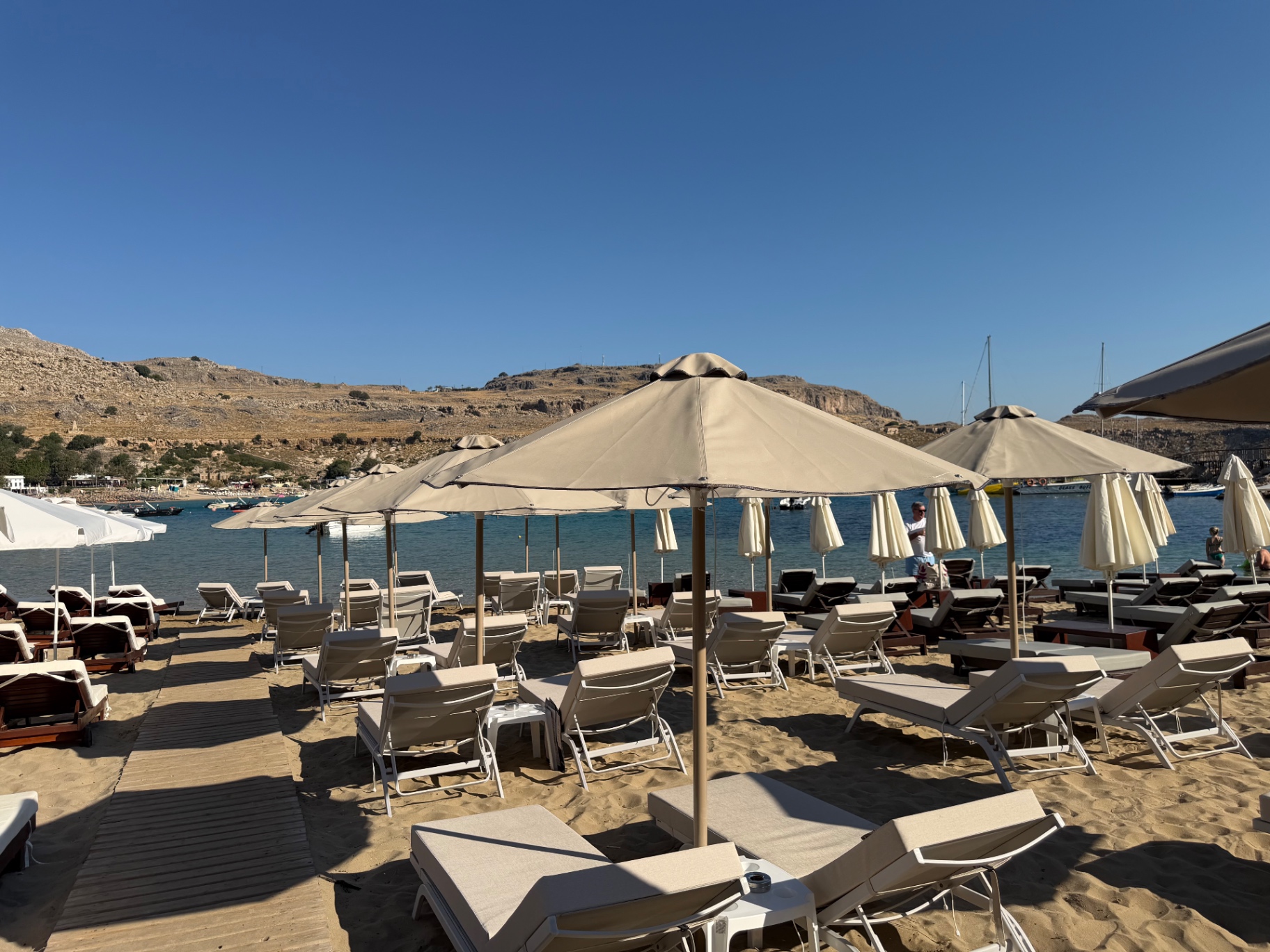 Pallas Beach in Lindos with beige parasols, sunbeds and the calm bay behind them